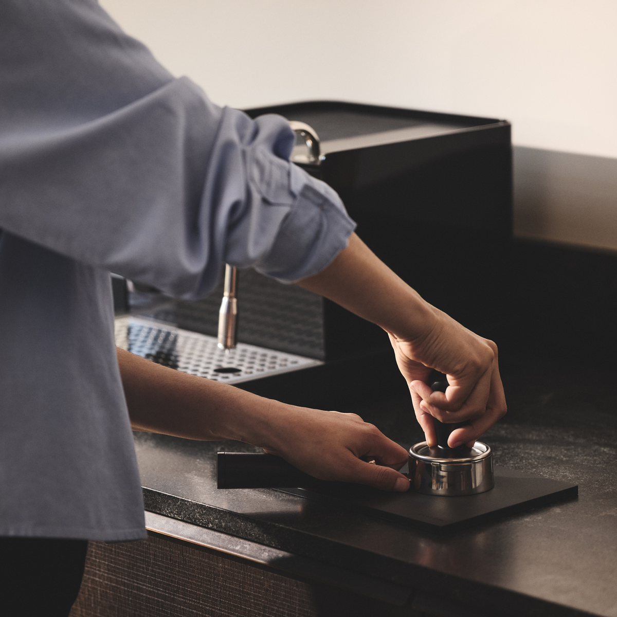 A person tamps coffee on a Fellow Espresso Tamping Mat beside an espresso machine.